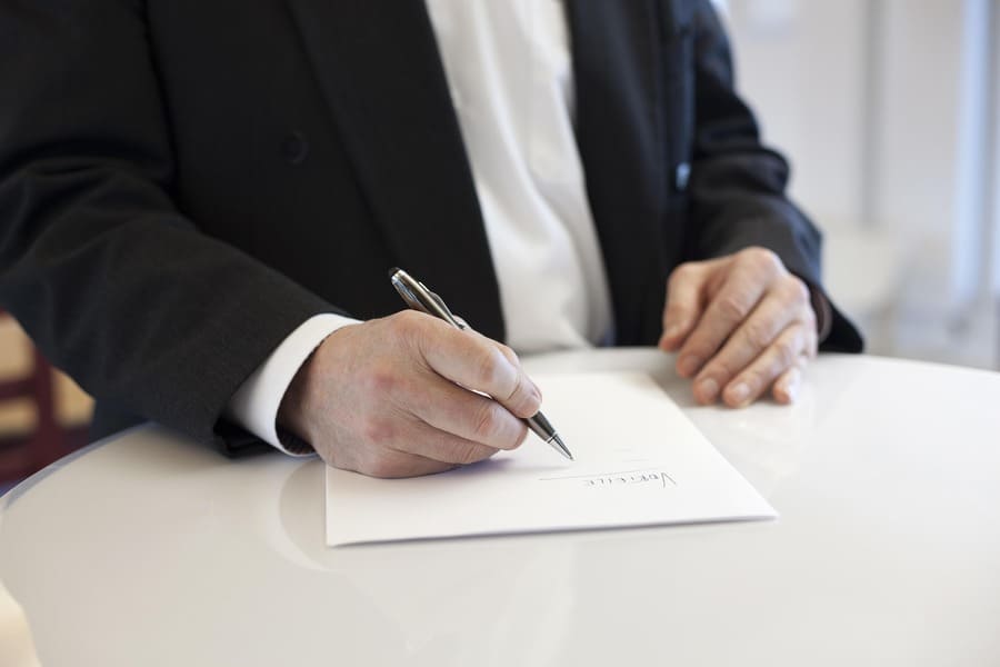 hombre con chaqueta azul marino firma documento sobre una mesa color beige claro