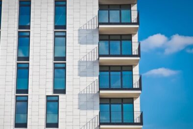 Edificio blanco con balcones con cristalera de tres piezas en tonos azules por el reflejo del cielo. Día despejado con cielo azul y pocas nubes